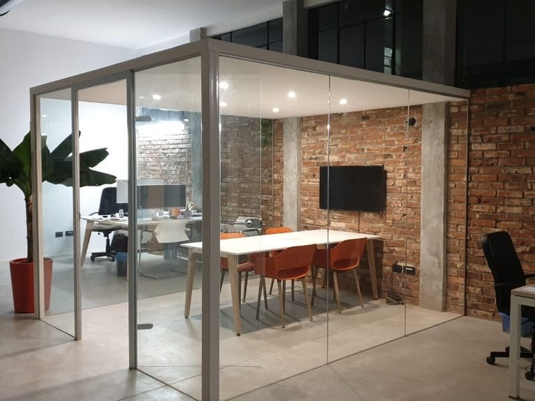 Modern glass-walled office meeting room with brick wall, conference table, orange chairs, and desk workspace inside an open industrial loft setting