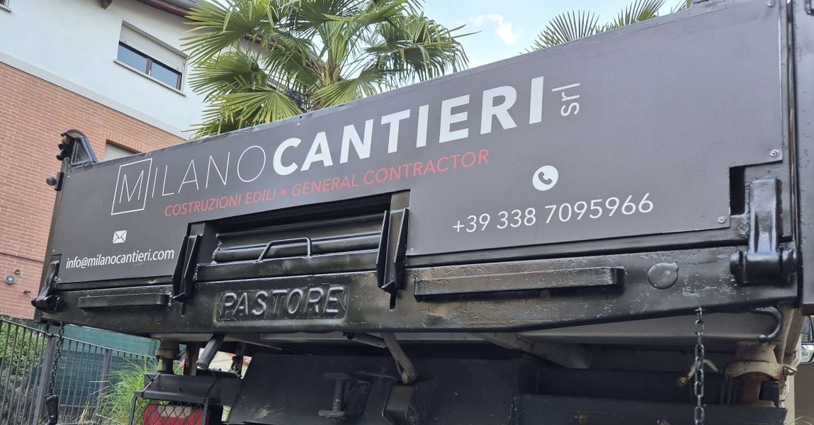 Gray construction truck with Milano Cantieri branding and contractor contact information parked outside a brick building