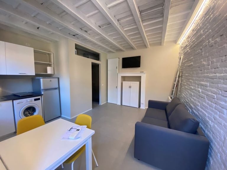 Modern studio apartment interior with open floor plan, dining table, kitchenette, blue sofa, exposed brick wall, and skylight ceiling